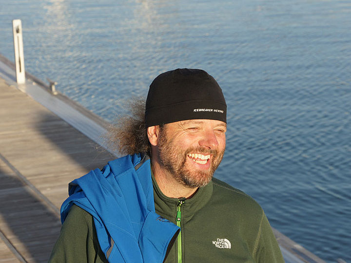 Ein Mann mit schwarzem Hut und grüner Jacke steht lächelnd auf einem Steg und ist bereit für seinen nächsten Einsatz.