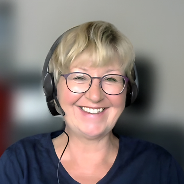 Eine lächelnde Frau mit Brille und Headset sitzt vor einem unscharfen Hintergrund mit dem Thema Mission.