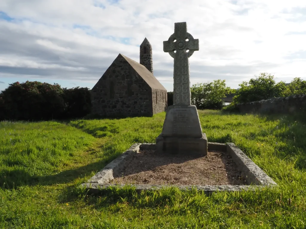 Ein steinerner Grabstein mit keltischem Kreuz steht vor einer alten Hebriden-Kirche und einem runden Turm auf einer Wiese.