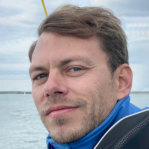 Ein Mann mit kurzen Haaren und blauer Jacke lächelt an einem bewölkten Tag am Wasser, bereit für seine Mission.