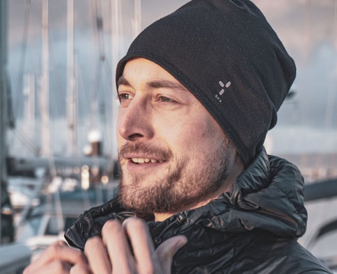 Mann mit schwarzer Mütze und Jacke lächelt im Freien in der Nähe von Booten, bereit für seinen nächsten Einsatz.